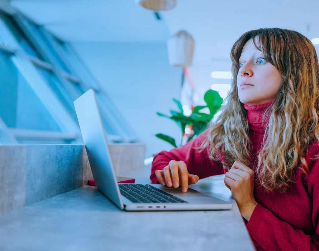 Woman at a laptop looking wide-eyed and tense, reacting to something unexpected on screen