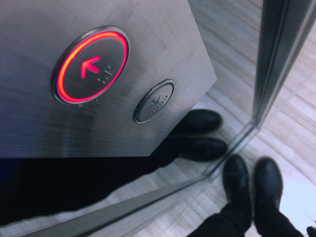 Close-up of a lit elevator button with a person's feet reflected below, suggesting upward movement and business scale