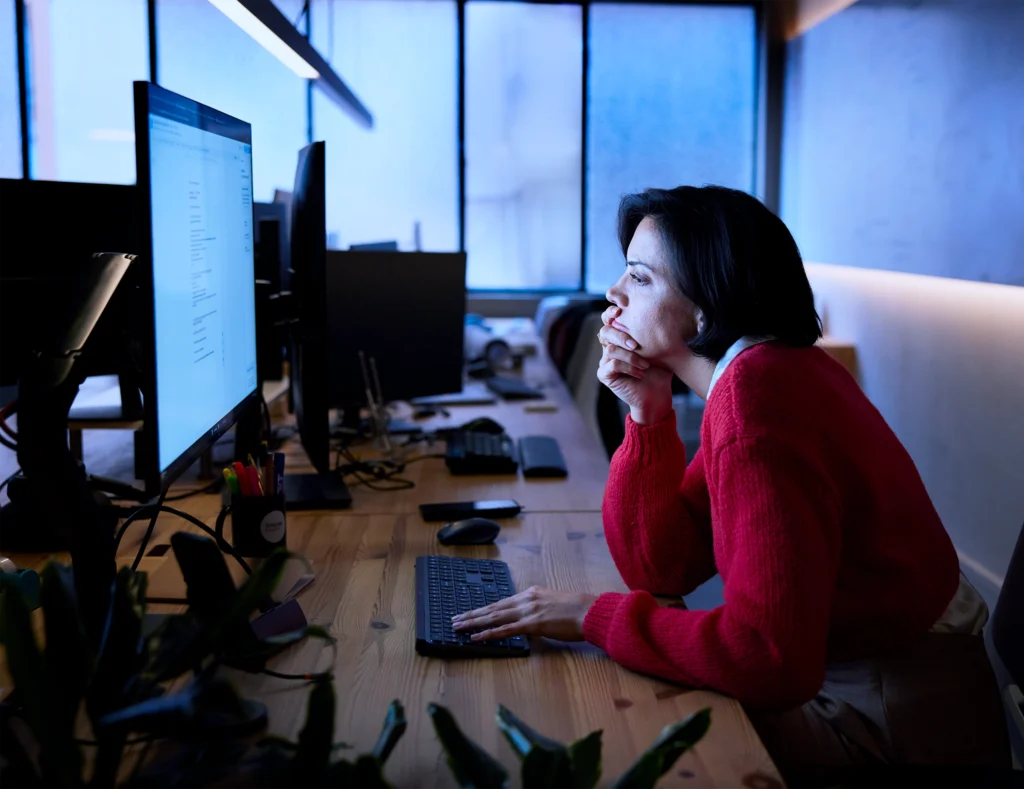 Alt text Travel manager analysing data on multiple screens in a dimly lit office, focused on programme performance