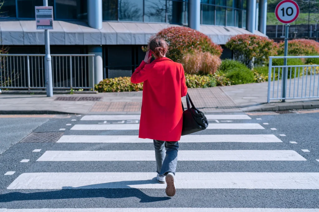 Business traveller in a red coat crossing a zebra crossing near an office building, travel bag in hand