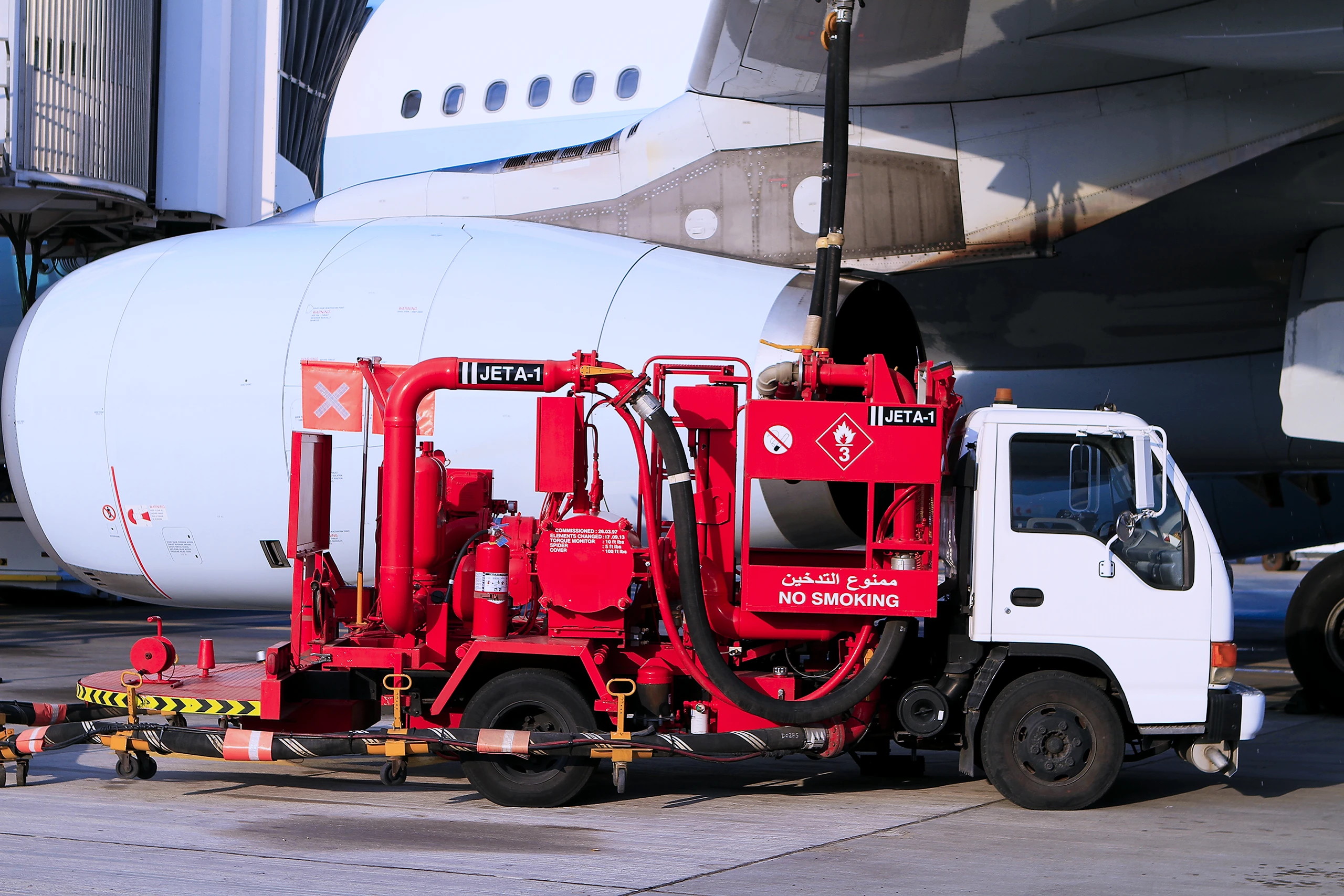A red Jet A-1 aviation fuel bowser truck refuelling a wide-body aircraft on an airport apron