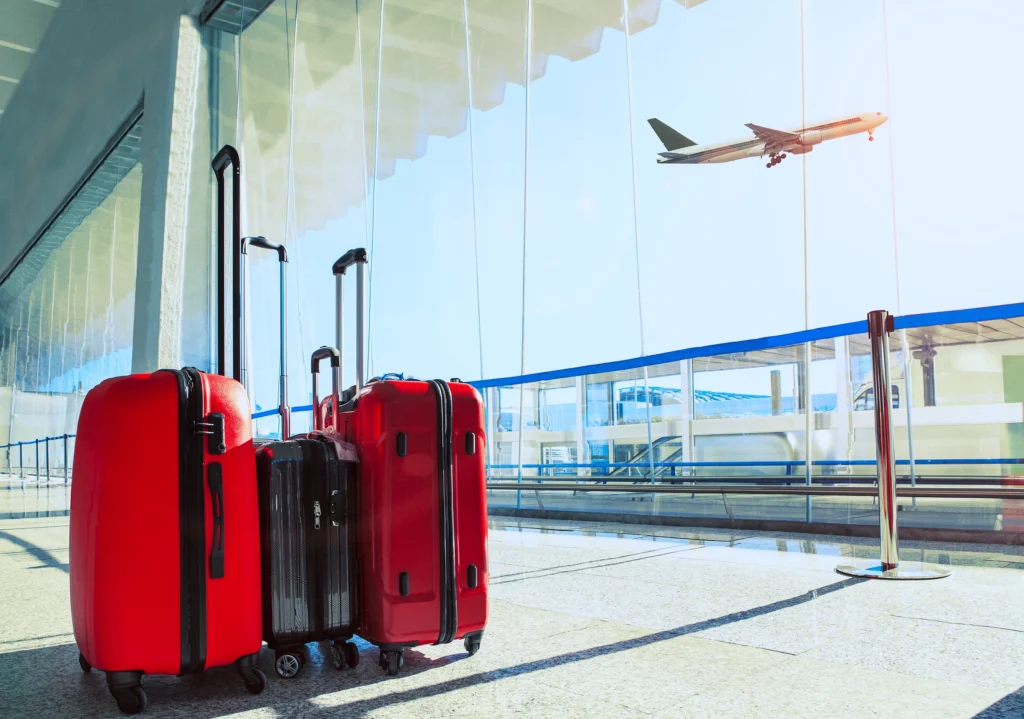 Red luggage standing in an airport terminal with a commercial aircraft visible through the glass facade in the background.